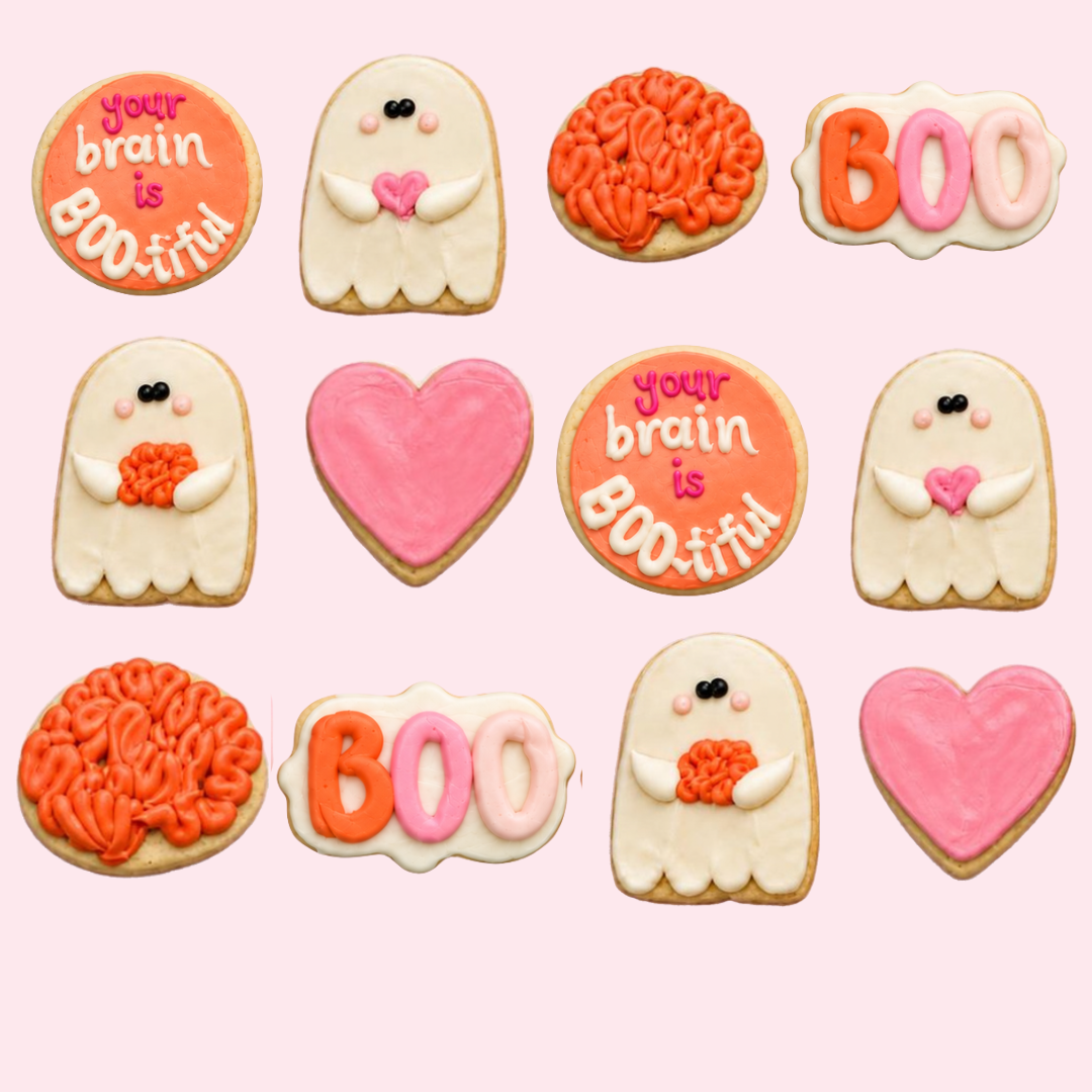 A set of Halloween-themed sugar cookies decorated with orange, pink, and white icing, arranged on a solid light pink background. The designs include ghost cookies with small pink hearts, brain-shaped cookies, a heart-shaped cookie, and a round cookie that reads 'your brain is BOO-tiful'.