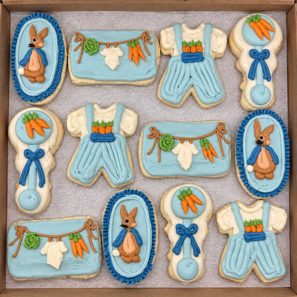 A set of baby shower themed sugar cookies decorated with light blue and white icing, featuring cartoon bunnies, carrots, and baby clothes, arranged in a square cardboard tray on a light gray surface.