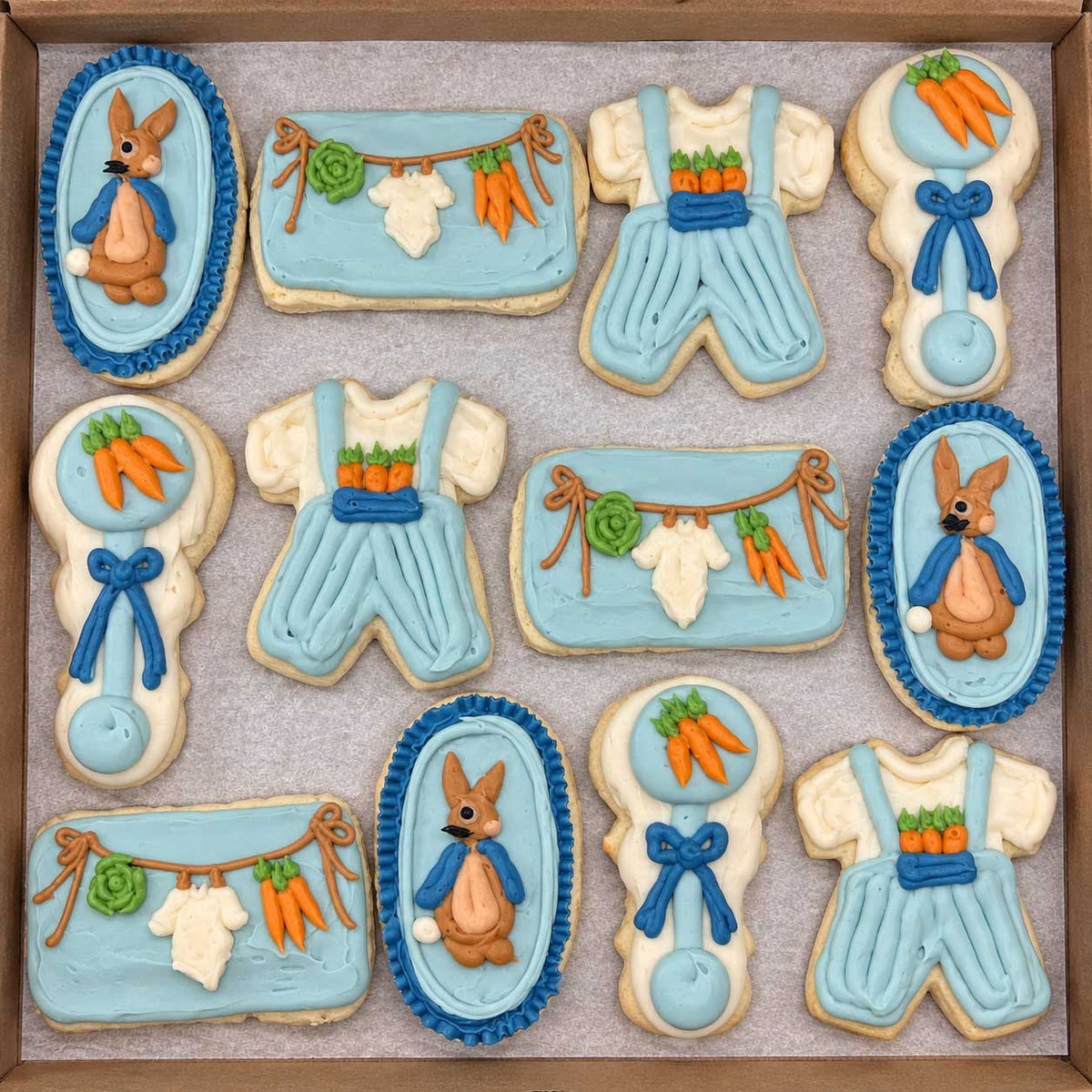 A set of baby shower themed sugar cookies decorated with light blue and white icing, featuring cartoon bunnies, carrots, and baby clothes, arranged in a square cardboard tray on a light gray surface.